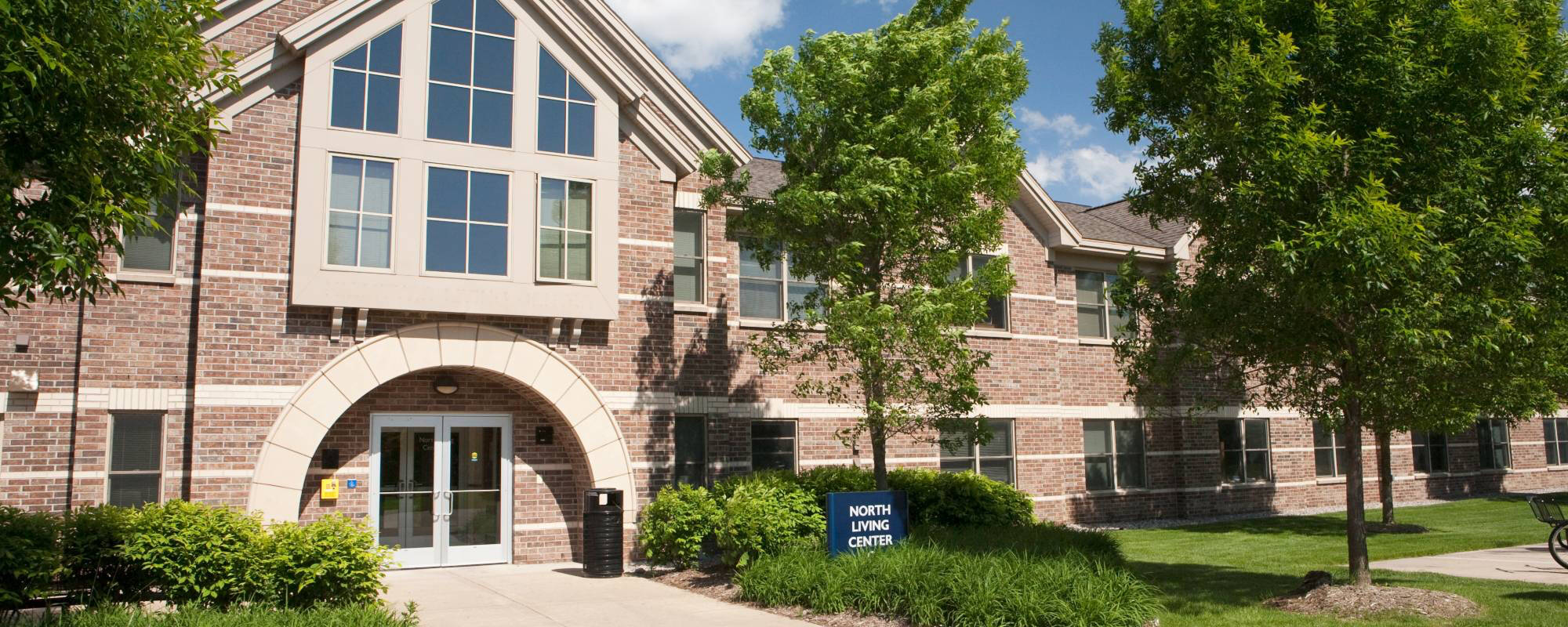 A two-story brick building with large windows and an arched entrance surrounded by green trees and bushes. A sign reads "North Living Center."